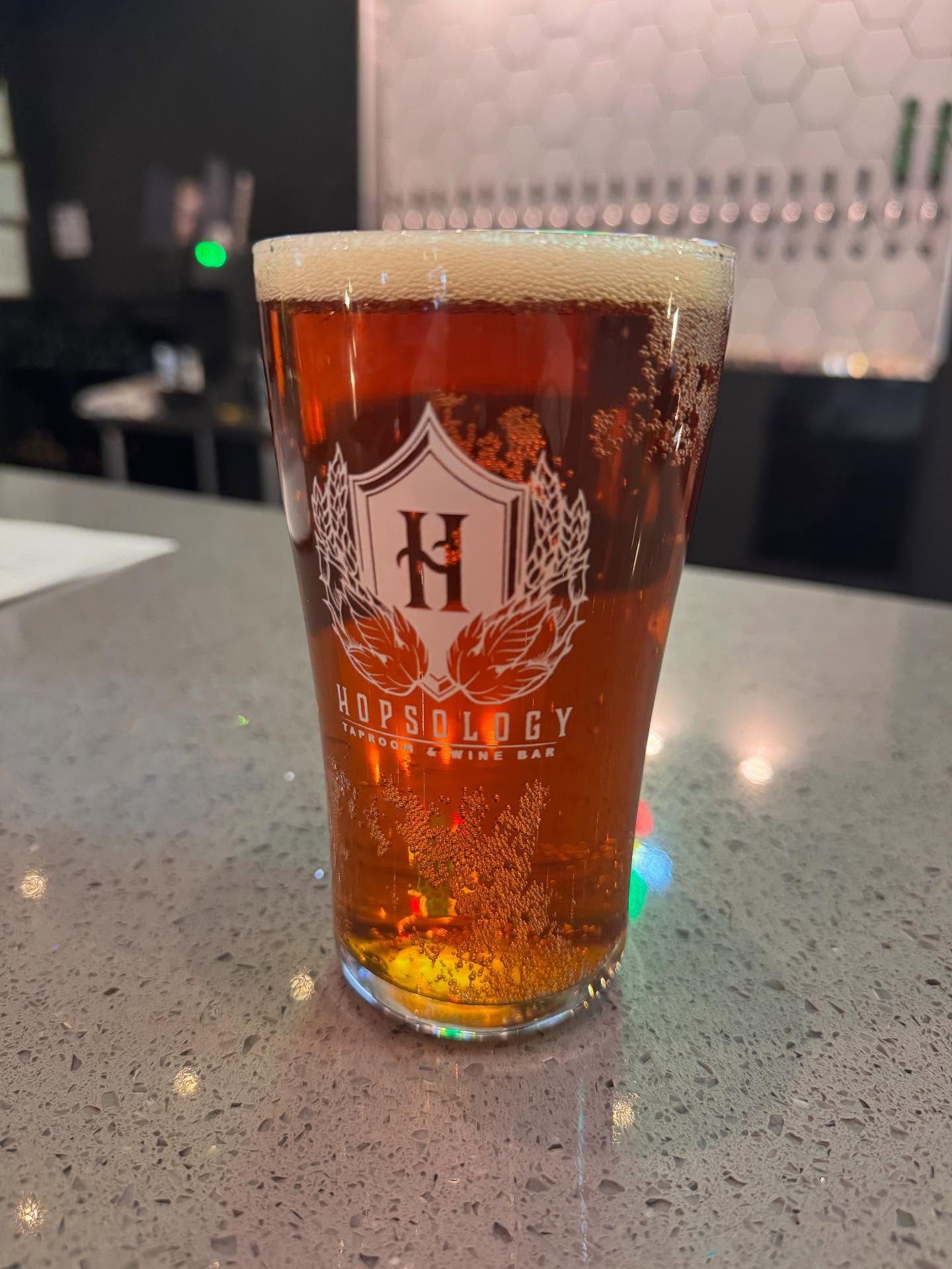 A Hopsology-etched pint glass of amber beer in front of the tap wall